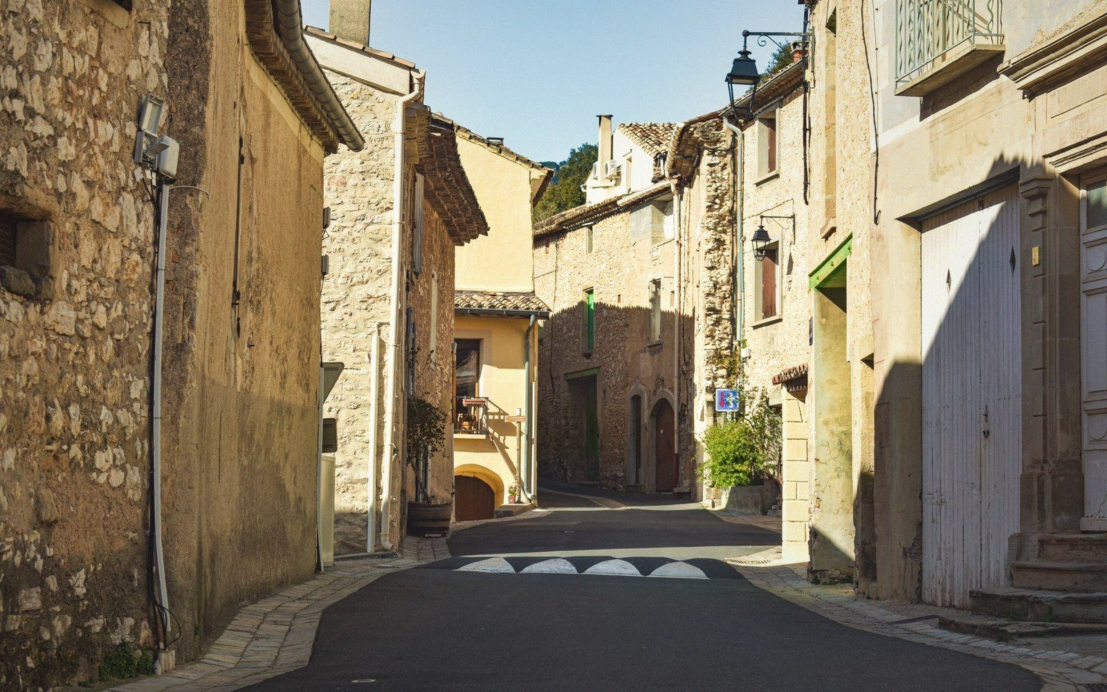 Narrow stone-walled street winding through a sunlit village with shuttered houses