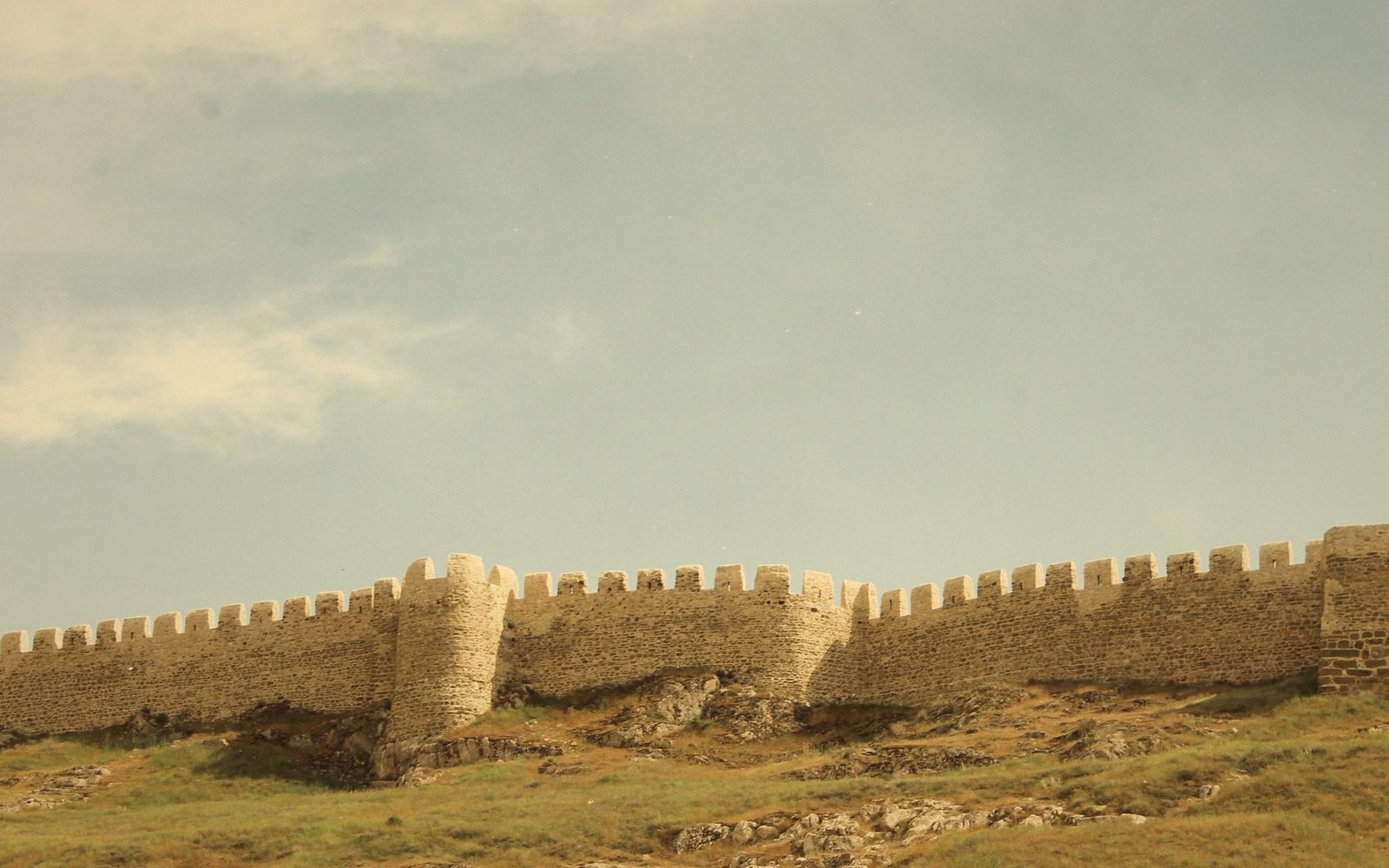 Crenellated stone fortress wall with a round tower atop a grassy hillside