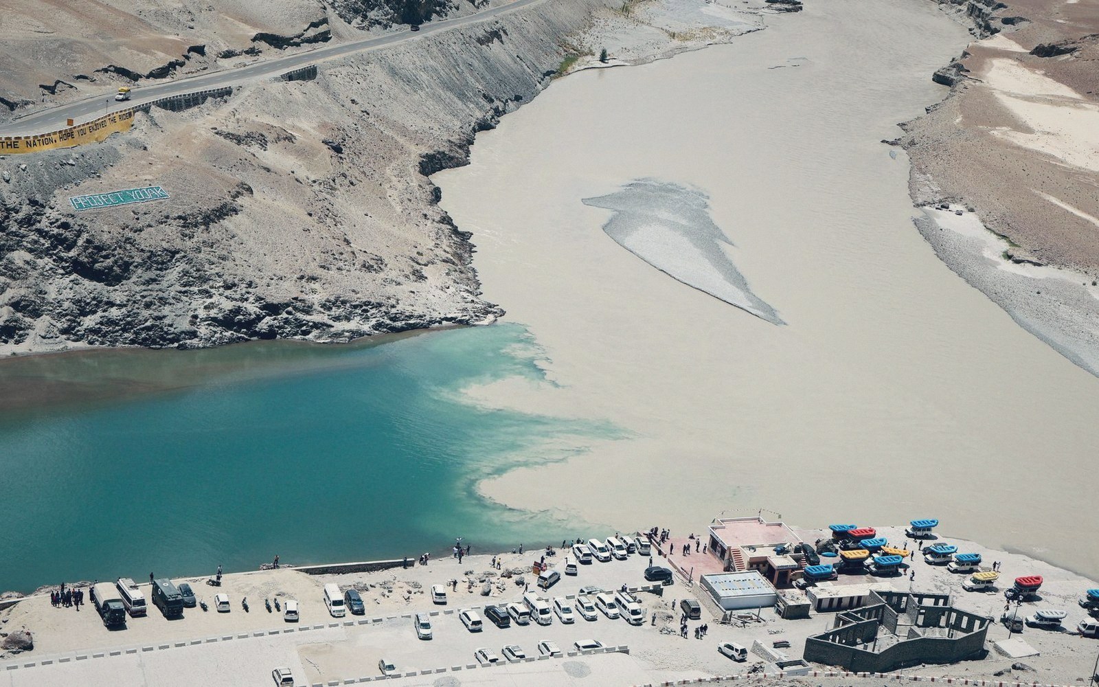 Two rivers of contrasting turquoise and silty grey water merge at a rocky confluence with parked vehicles nearby