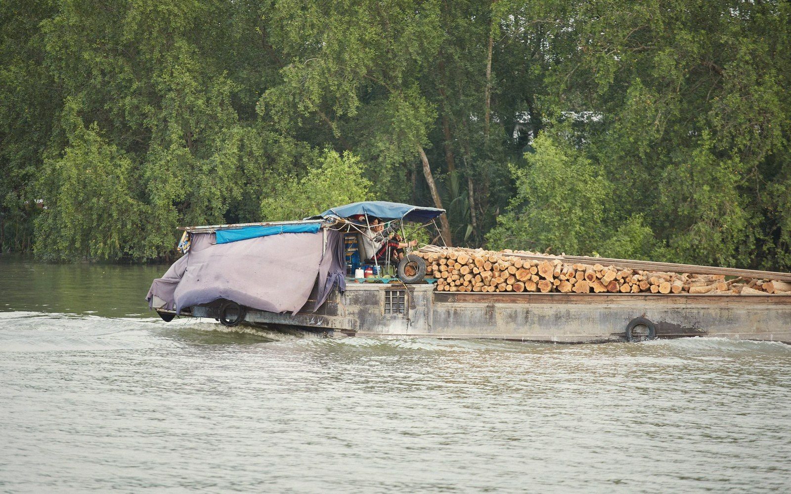 A tarp-covered river barge stacked with cut logs travels along a tree-lined waterway