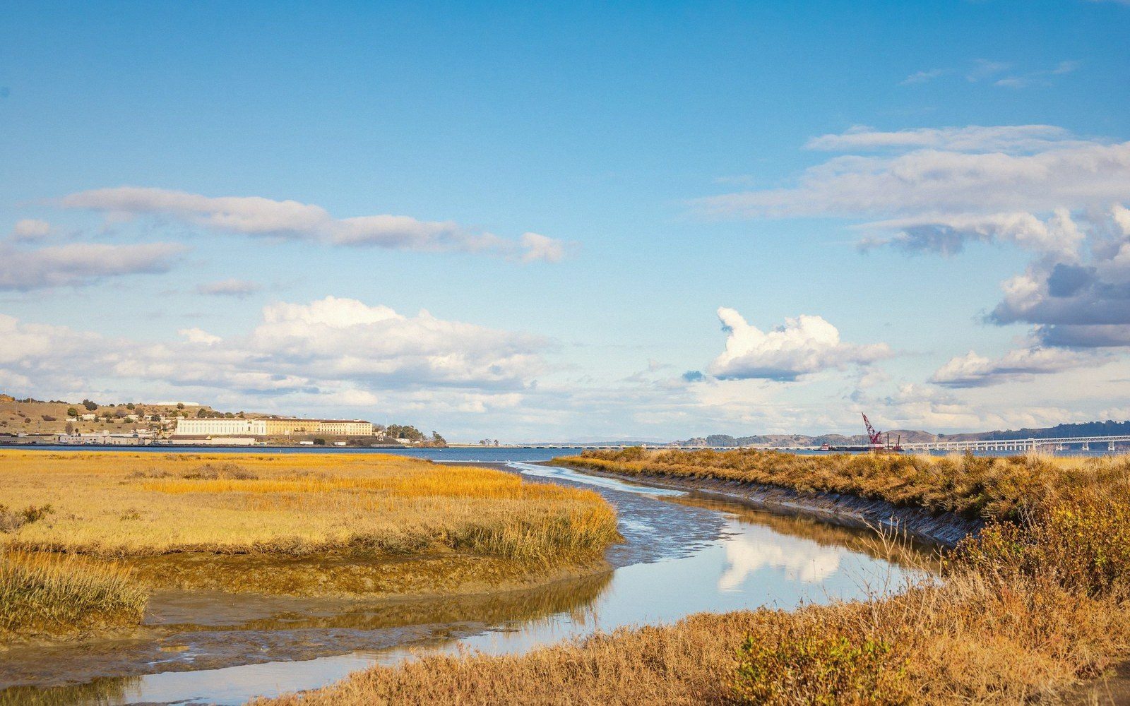 A tidal creek winds through golden marsh grasses toward a distant waterfront town under a partly cloudy sky