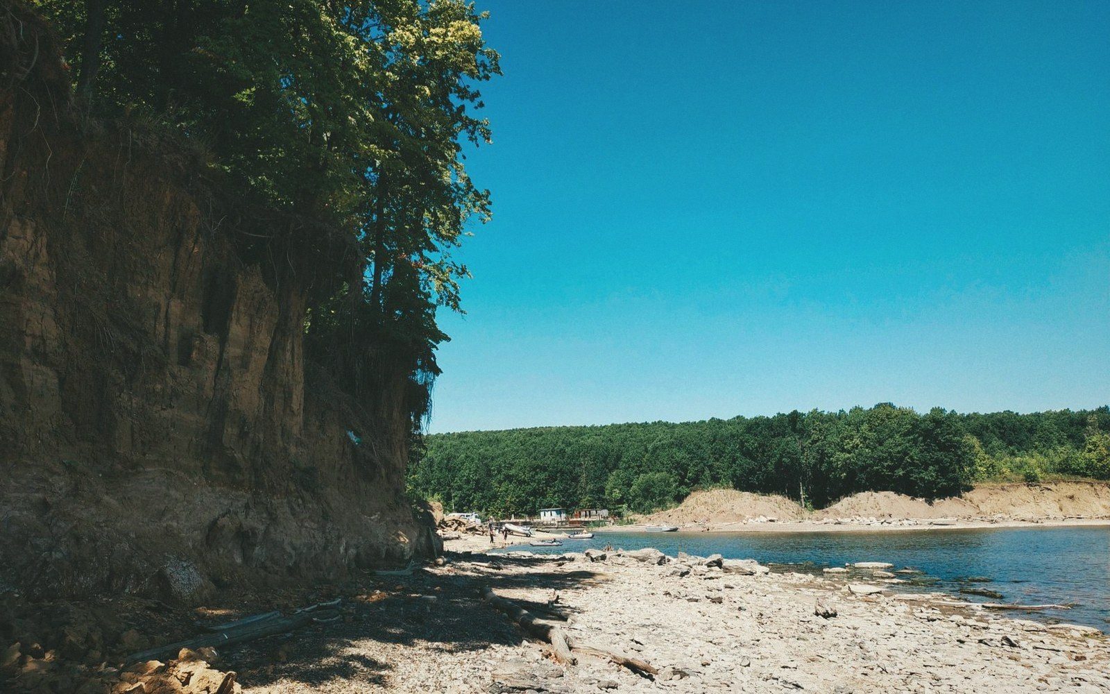 A tree-topped cliff rises above a rocky riverbank under a clear blue sky
